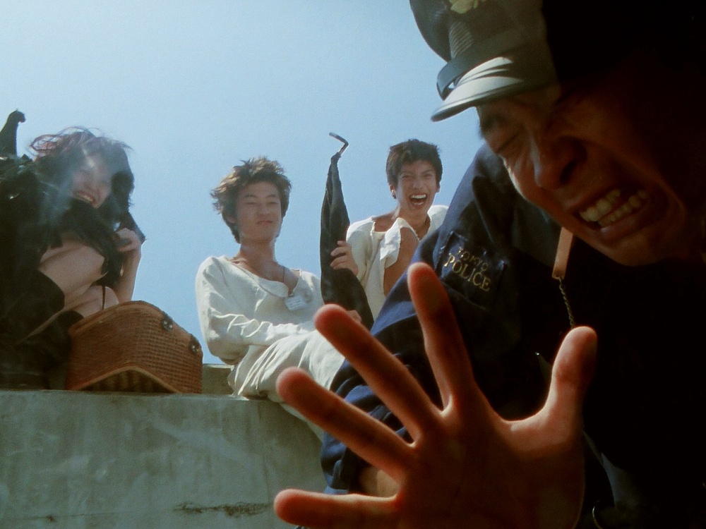 Three young Japanese people in loose clothing laugh and stare at a man whose face is squashed against the camera