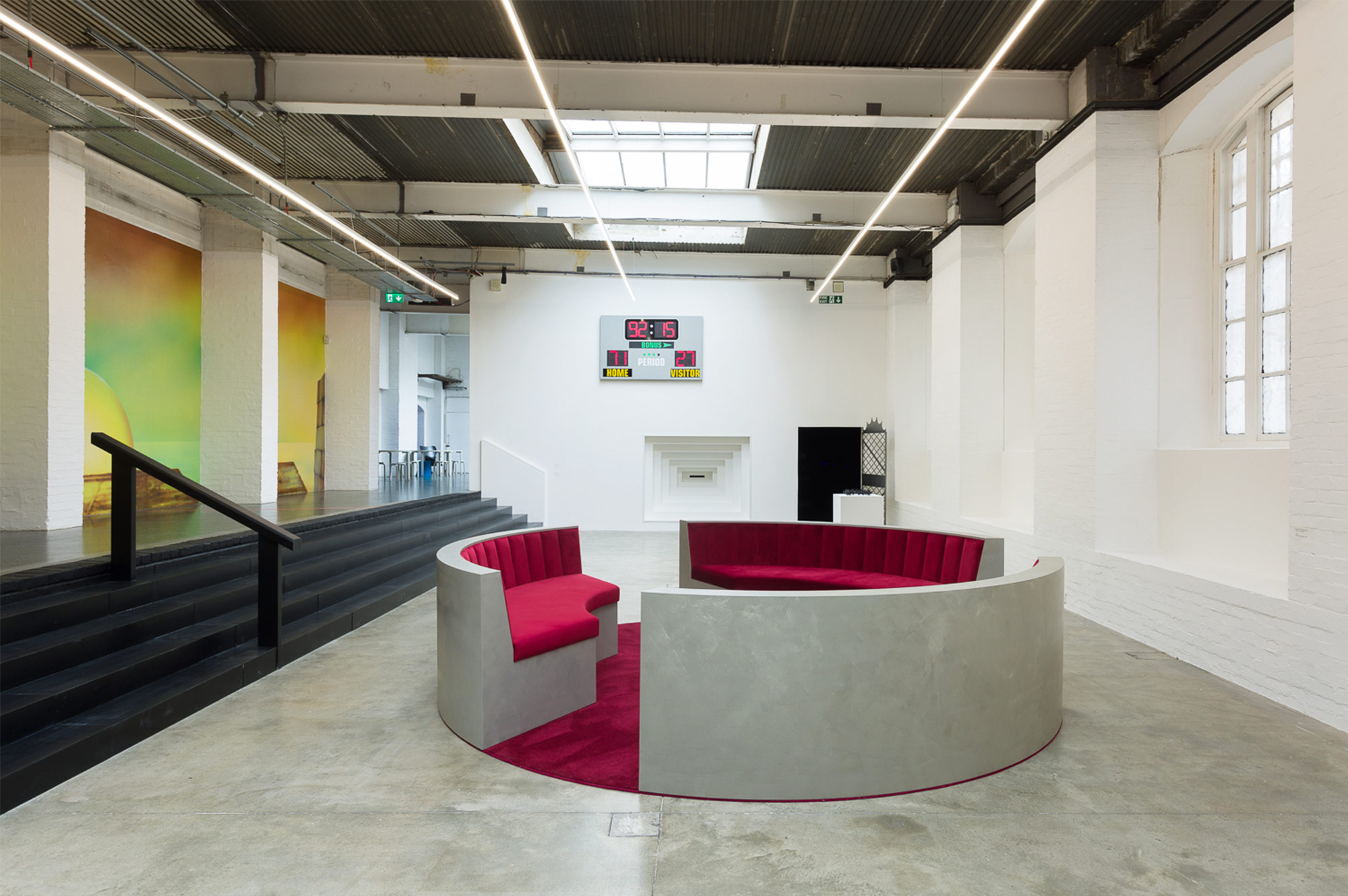 The main gallery for The Red Sun Is High, The Blue Low. On the left wall, painted stills of sunsets. Black stairs lead down to the gallery floor, where three sofas sit in a circle, with plush red velvet covering and floor. On the back wall is a baseball-like scoreboard above a black doorway and a peephole.