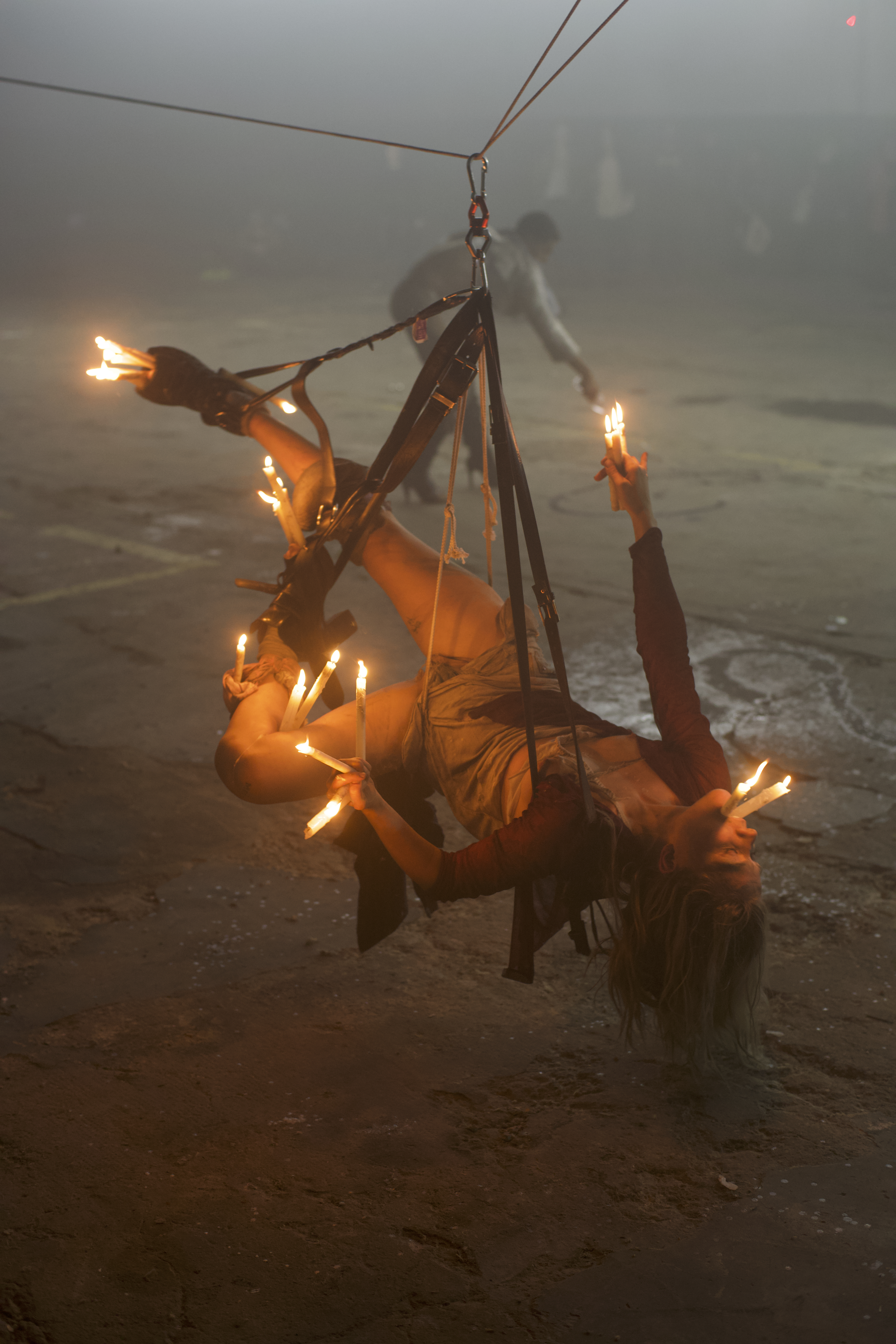 Maria Metsalu hangs from belts above a sand floor, holding candles in her mouth, fingers, and around her legs